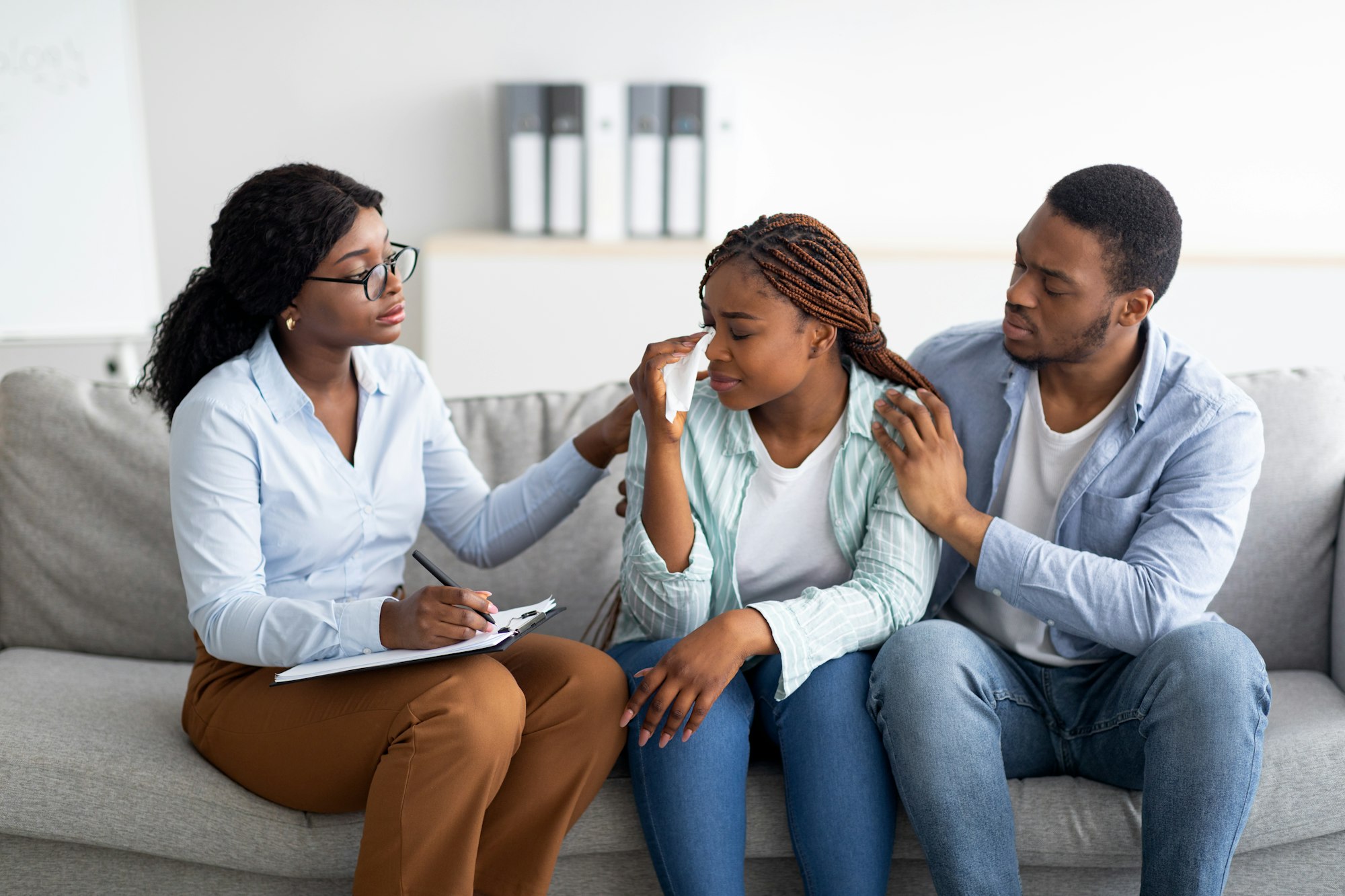 Millennial black woman crying at psychologist's office, loving husband supporting her with